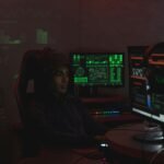 A man in a black hoodie engaged in cybersecurity work using multiple monitors indoors.