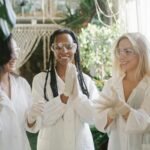 Three diverse women scientists smiling in a lab with plants, wearing lab coats and goggles.